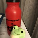 Red bottle printed with "Malice Domestic" and cup of poisoned tea; cute green frog stuffed animal in front of bottle