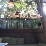 Image of patio with wooden bench and cushions plus hanging lights strung over a tree